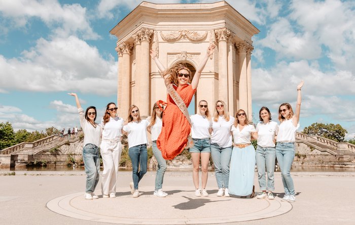 Groupe de femmes célébrant un enterrement de vie de jeune fille à Montpellier, posant devant un monument emblématique avec des tenues coordonnées, dans une ambiance joyeuse et ensoleillée.