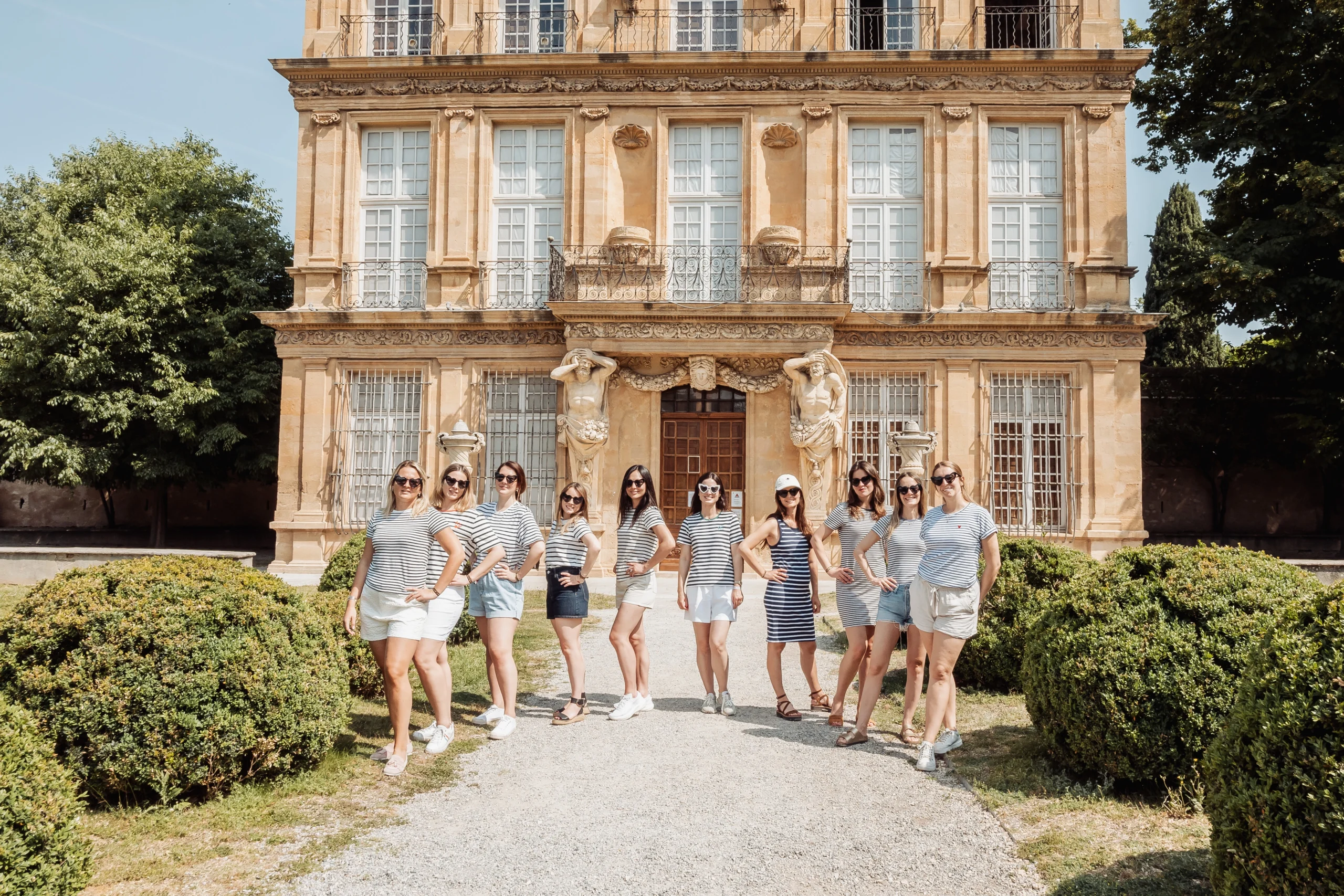 evjf-aix-en-provence-photo-groupe-rayures-devant-batiment Groupe de femmes en tenue marinière posant en ligne devant un bâtiment historique à Aix-en-Provence pendant un EVJF.