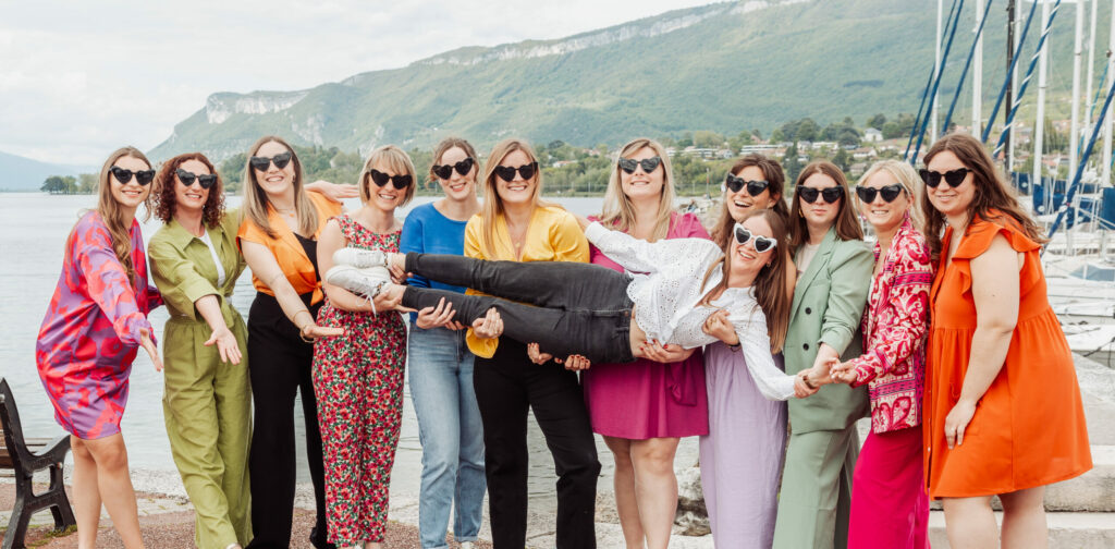 Photo EVJF à Aix-les-Bains avec un groupe d’amies qui soulèvent la future mariée en riant, au bord du lac du Bourget, toutes vêtues de tenues colorées et de lunettes en cœur