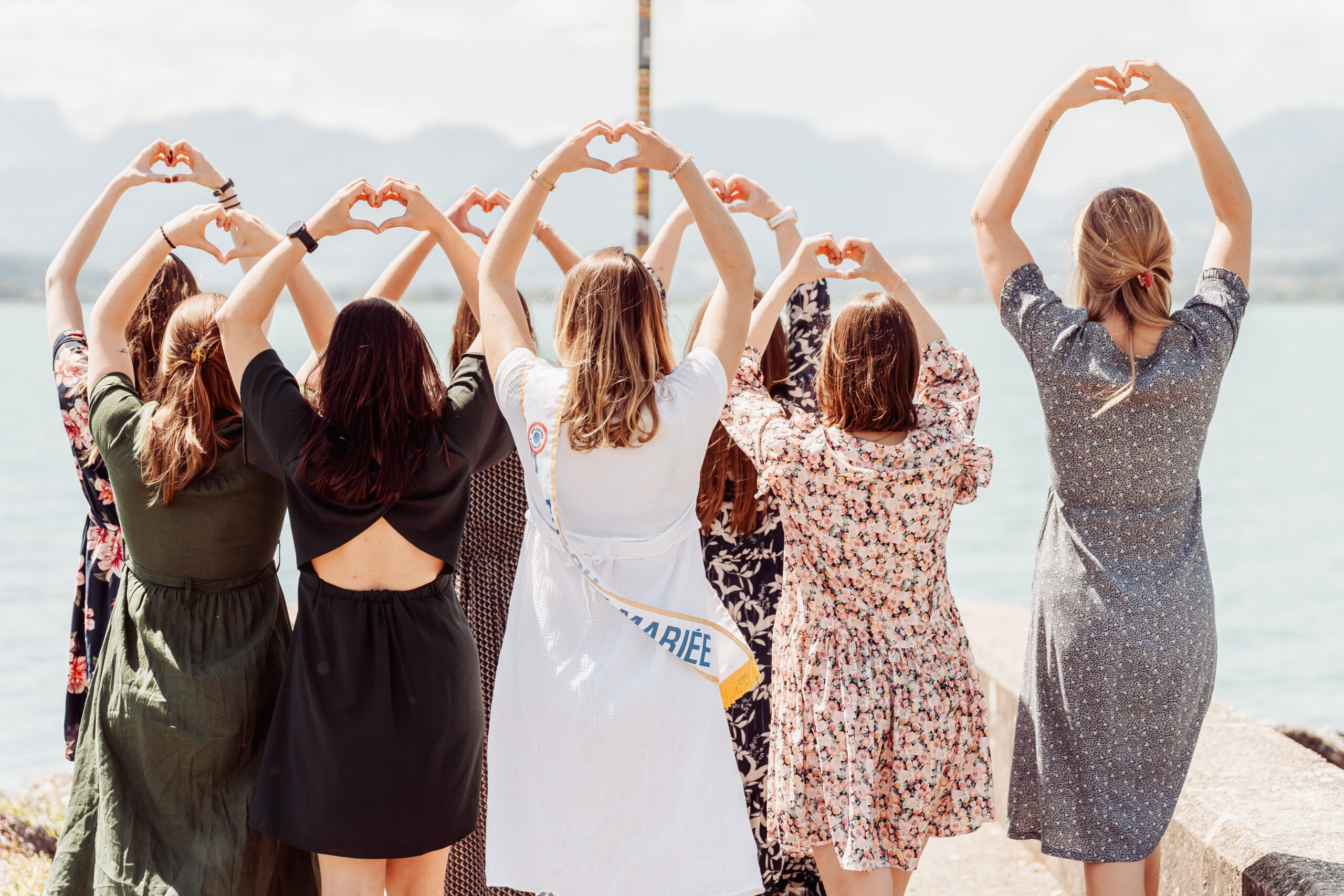 Photo de dos d’un groupe de femmes célébrant un EVJF à Aix-les-Bains, formant des cœurs avec leurs mains face au lac et aux montagnes, dans une mise en scène poétique et complice.
