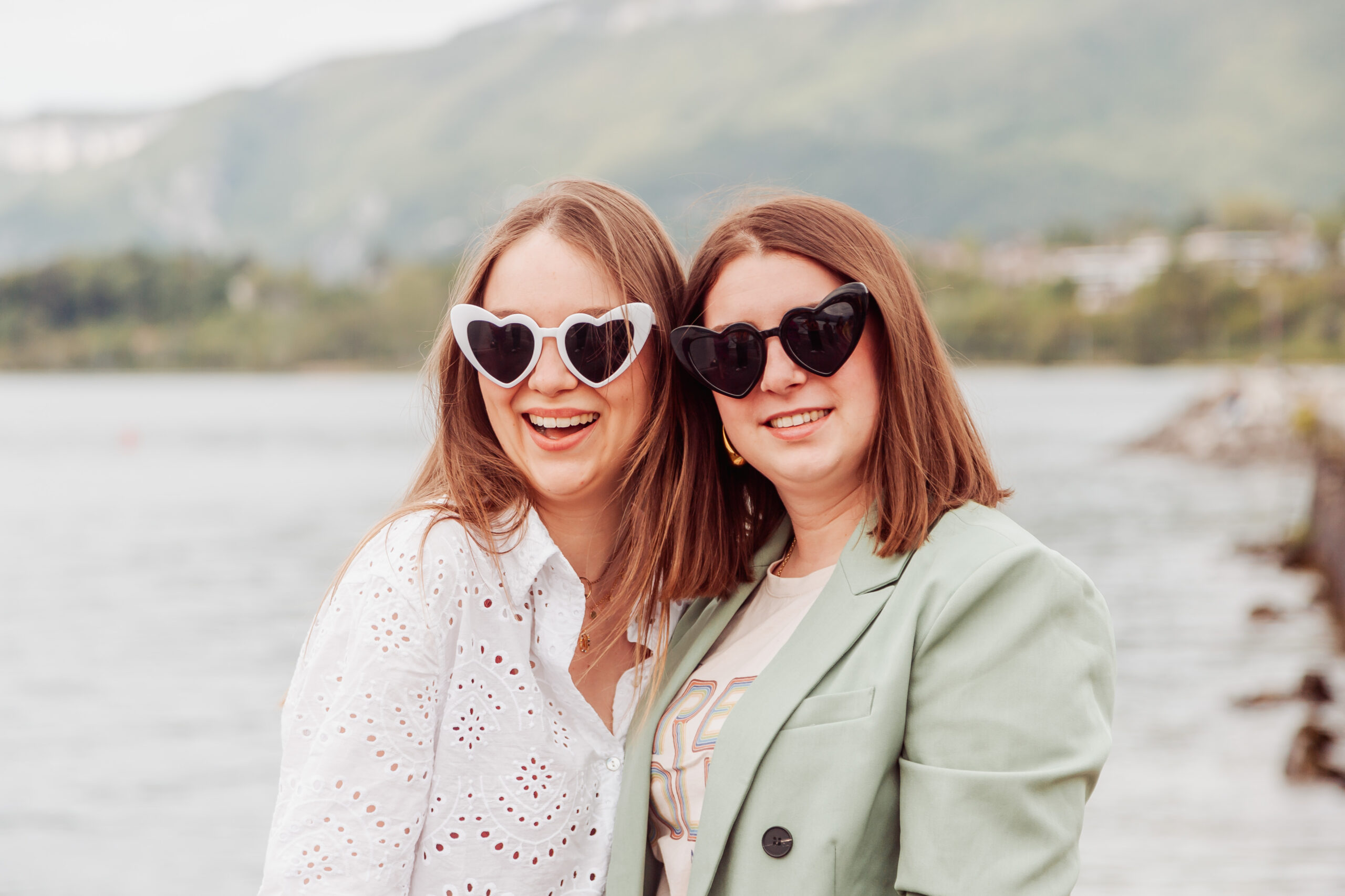 Portrait de deux amies lors d’un EVJF à Aix-les-Bains, souriantes et portant des lunettes en forme de cœur, posant devant le lac avec les montagnes en arrière-plan.