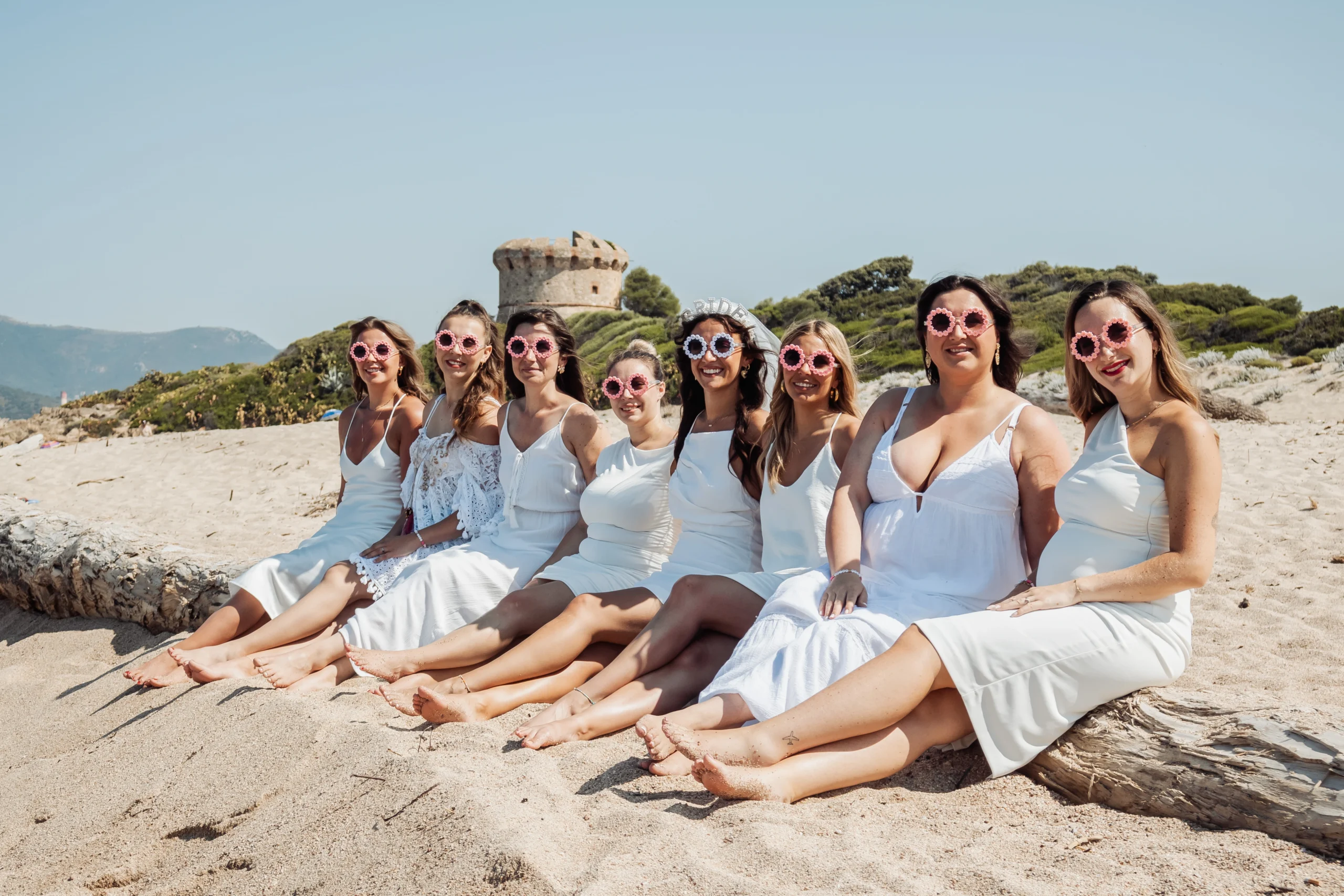 Groupe de femmes célébrant un EVJF à Ajaccio, assises en ligne sur la plage en robe blanche avec des lunettes roses fleuries, dans un décor naturel avec vue sur la montagne et une tour génoise.