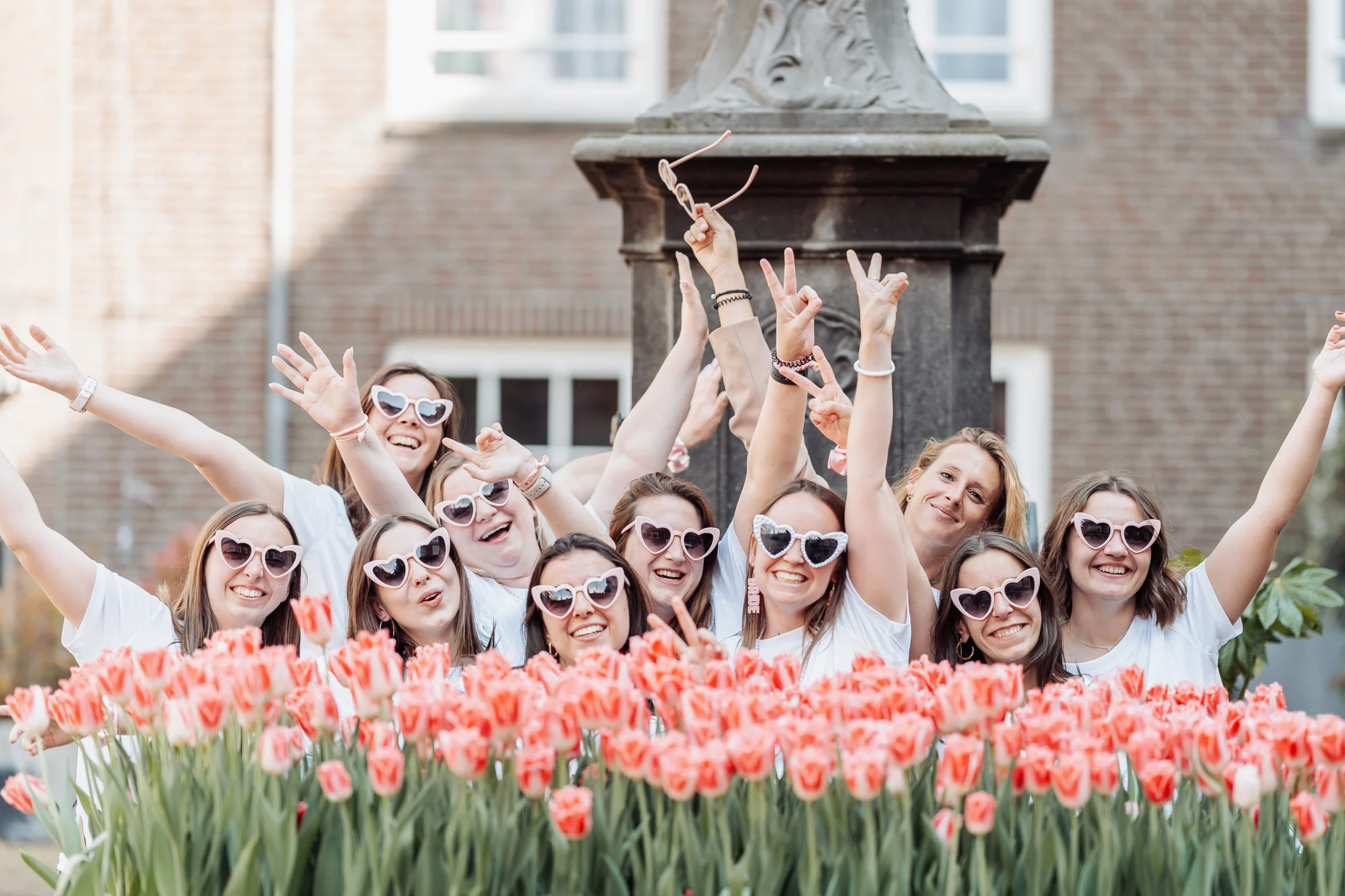 team-bride-amsterdam-tulipes-style-festif Photo de groupe d’un enterrement de vie de jeune fille à Amsterdam : des femmes souriantes portant des lunettes en forme de cœur posent derrière un parterre de tulipes roses, dans une ambiance joyeuse et printanière.
