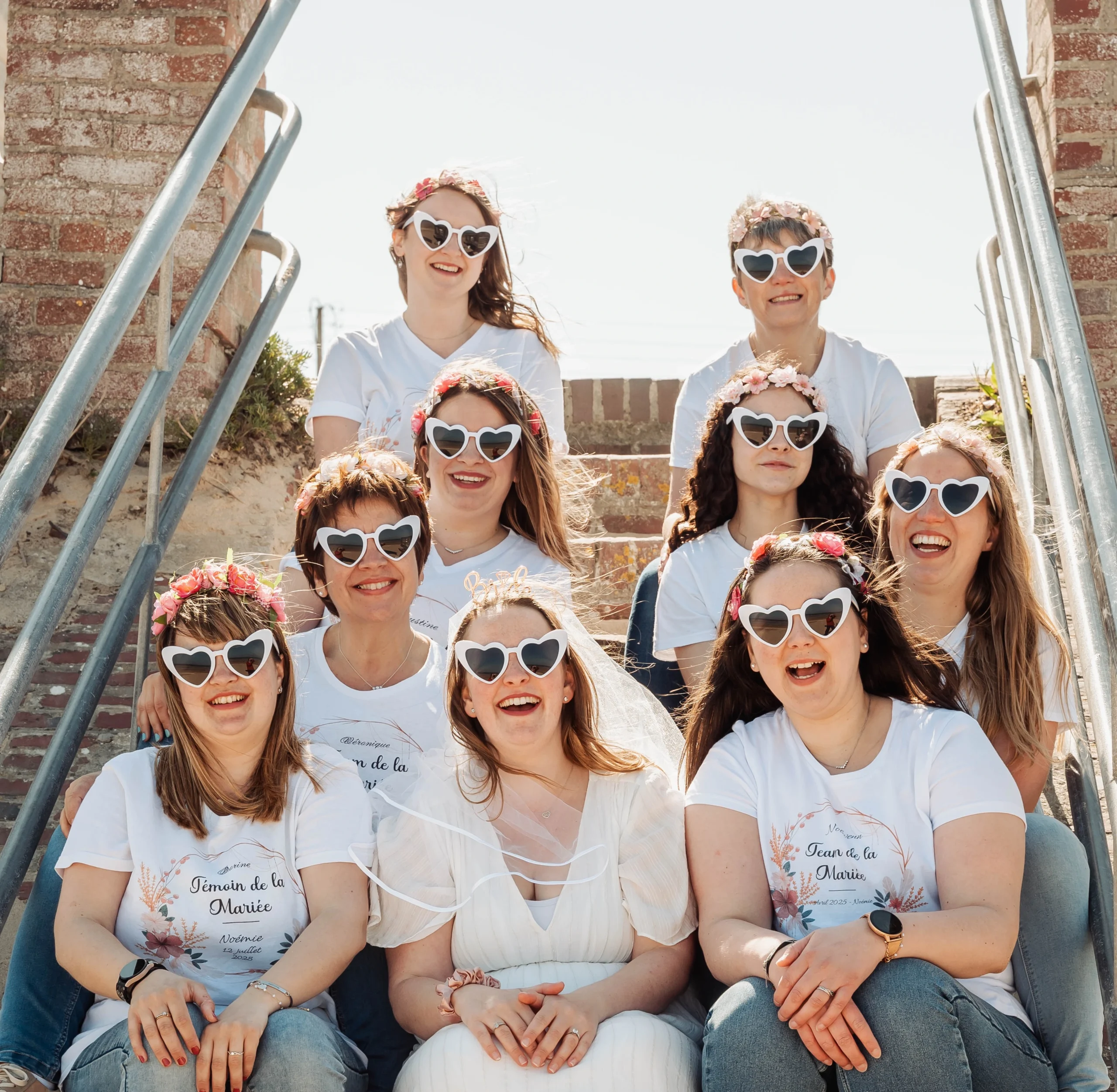 Groupe de femmes en EVJF à Cabourg, assises sur un escalier de plage ensoleillée, portant des lunettes blanches en forme de cœur.