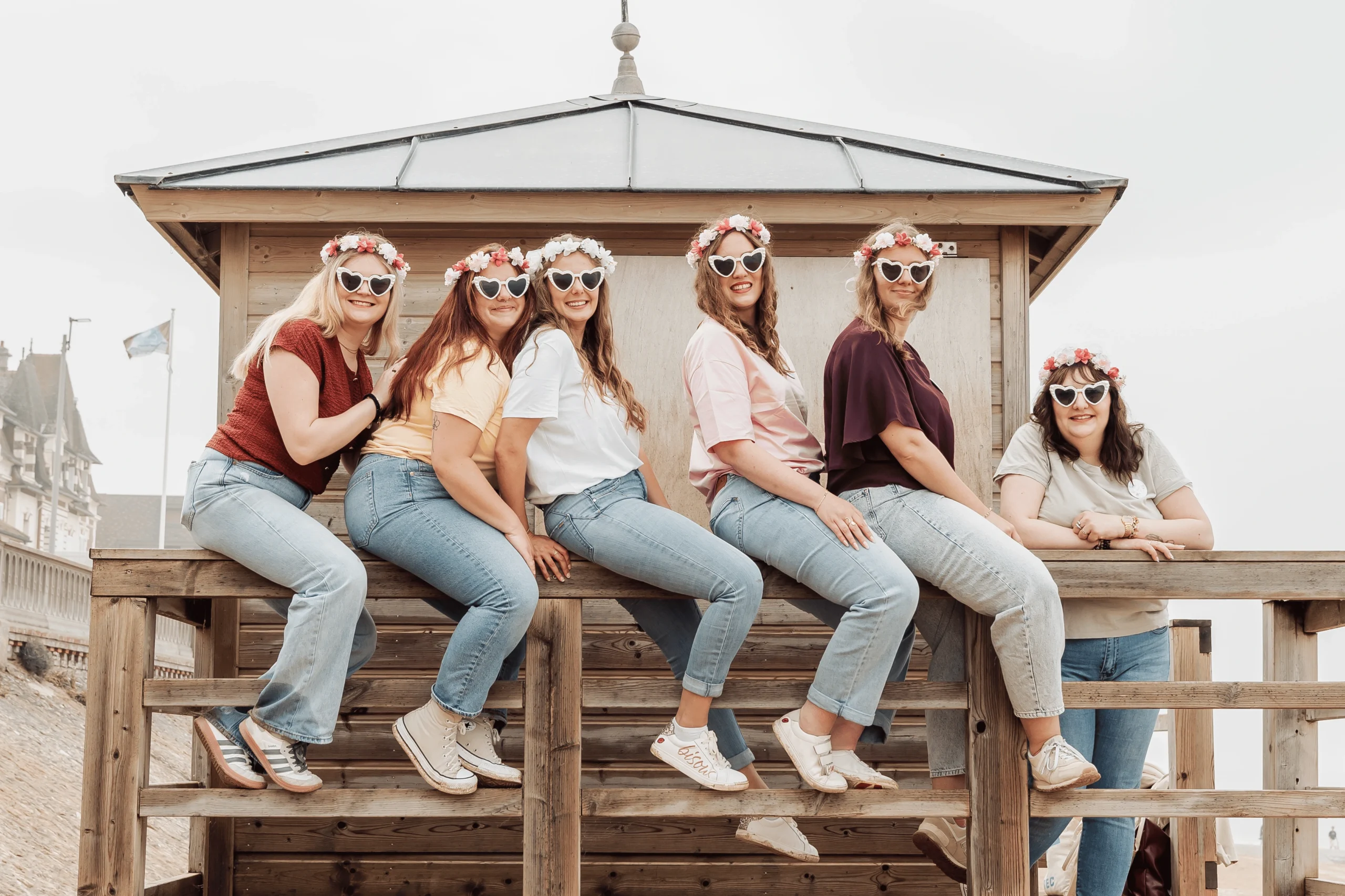evjf-cabourg-plage-cabane-photo-groupe Photo EVJF à Cabourg avec le groupe assis sur une cabane en bois face à la plage, lunettes cœur et couronnes de fleurs. Ambiance bord de mer et fun.