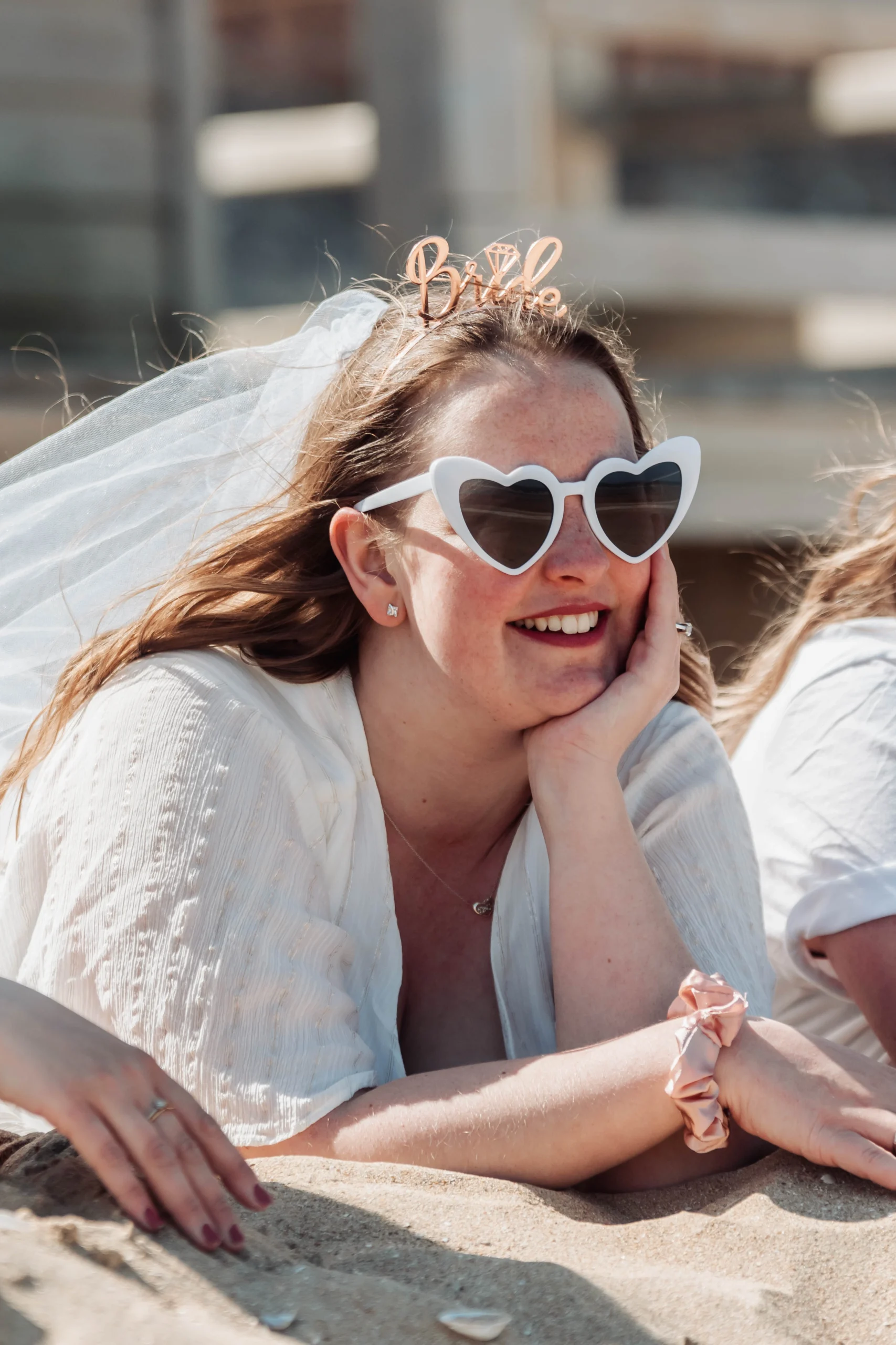 evjf-cabourg-portrait-mariee-sable-lunettes-blanches Portrait de la mariée allongée sur le sable à Cabourg lors de son EVJF, portant des lunettes blanches en forme de cœur et un accessoire "bride".