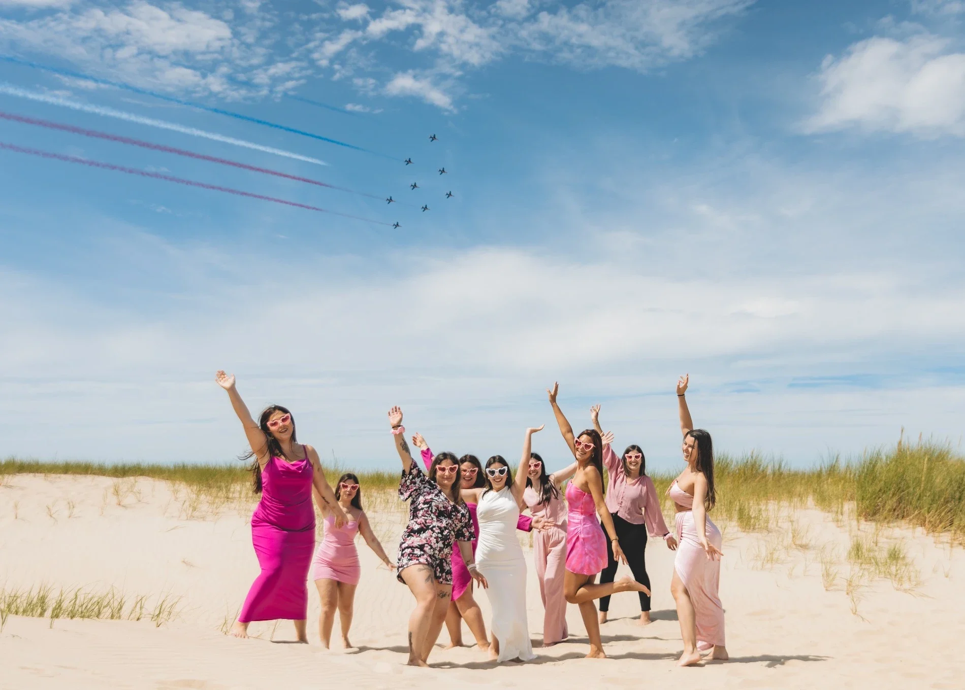 Groupe de femmes célébrant un enterrement de vie de jeune fille sur la plage du Touquet, toutes vêtues de tenues roses sous un ciel bleu, en train de lever les bras en riant.
