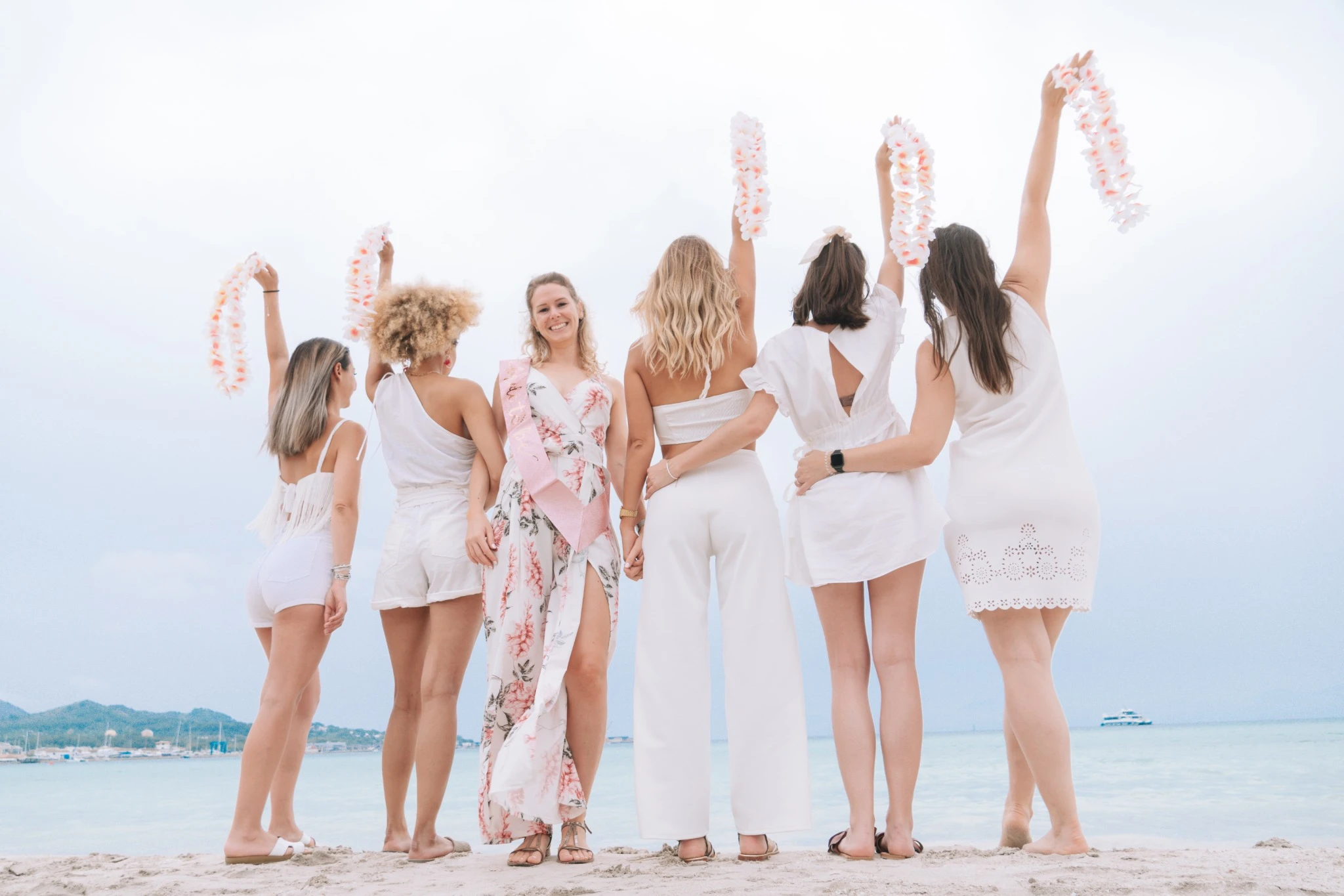 evjf-majorque-filles-de-dos-levees-colliers Filles de dos sur la plage de Majorque levant les colliers à fleurs pour célébrer l’EVJF