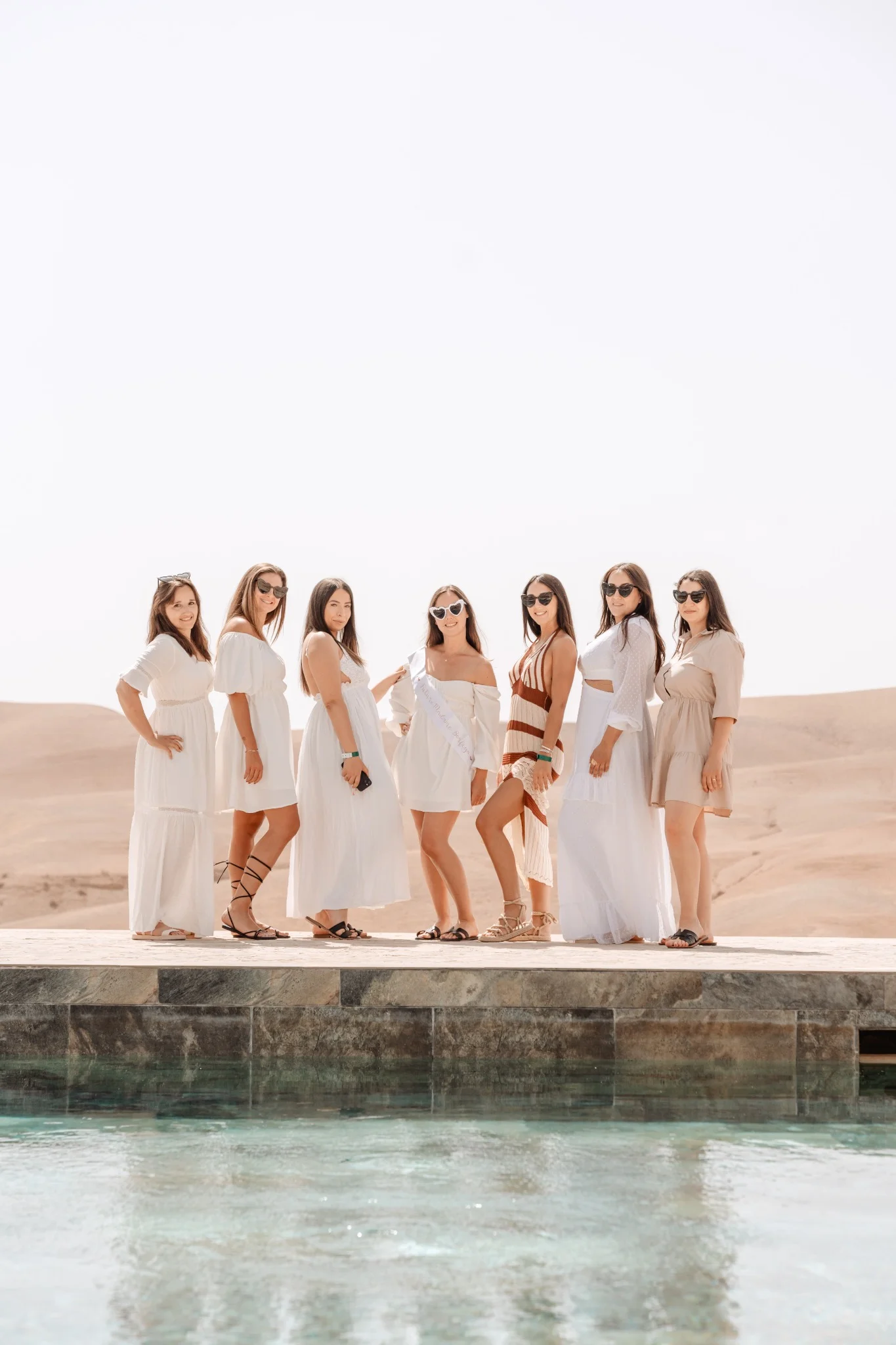 Groupe de femmes en tenues claires posant au bord d'une piscine dans le désert marocain lors d'un EVJF chic et élégant.
