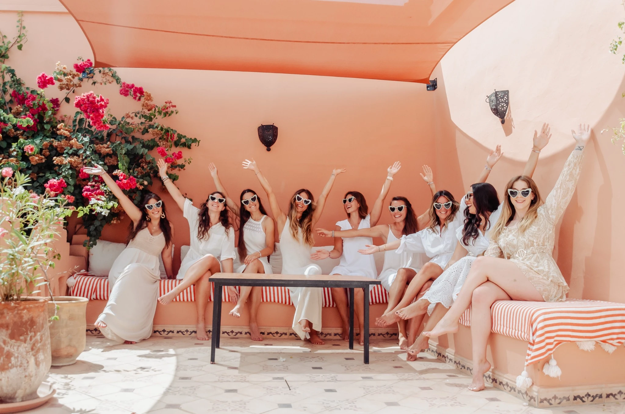 evjf-marrakech-amies-lunettes-terrasse Groupe de femmes en blanc avec lunettes cœur lors d’un EVJF à Marrakech sur une terrasse fleurie