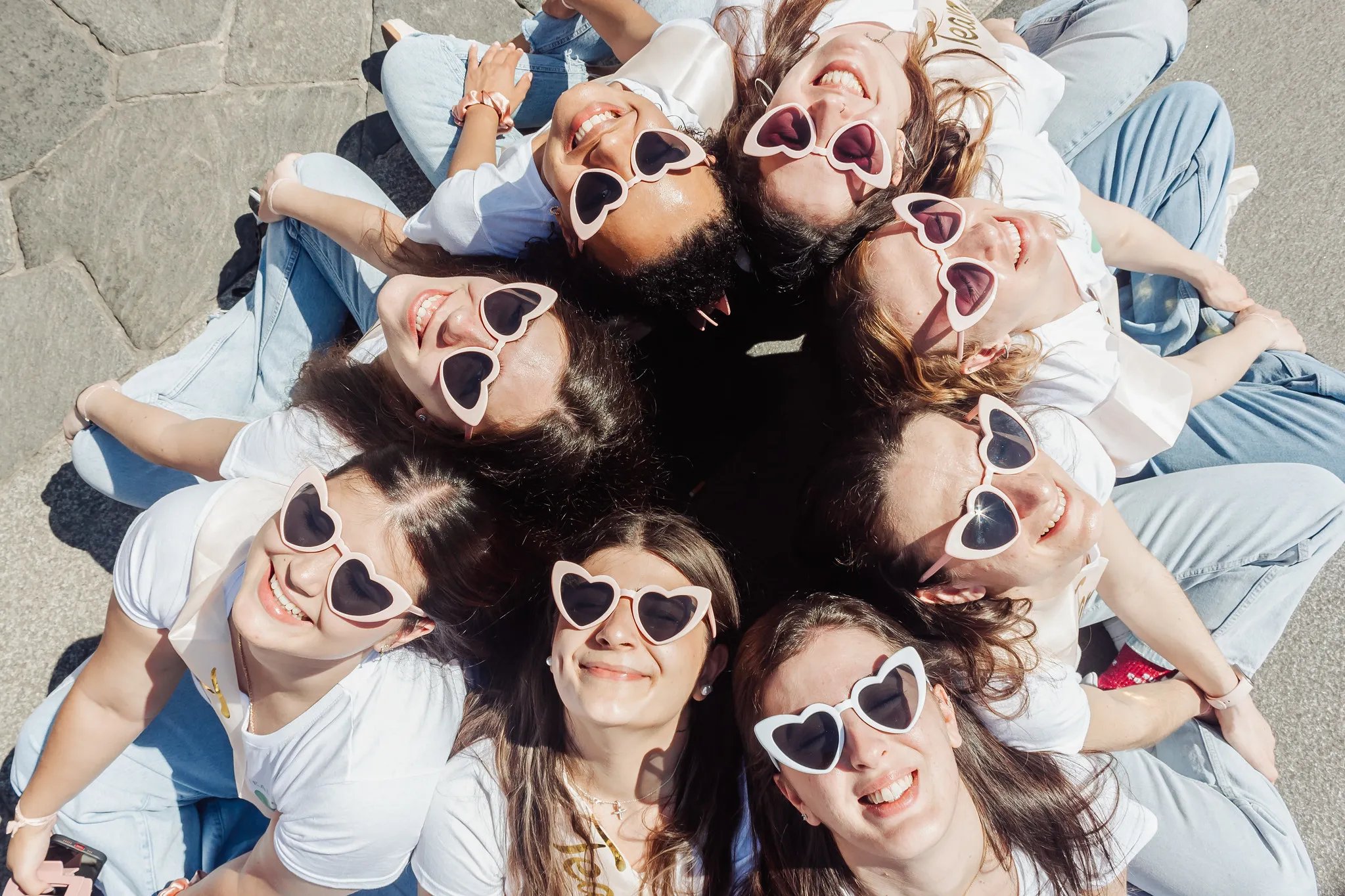 Photo de groupe EVJF à Milan avec lunettes en cœur et ambiance festive Groupe de femmes assises en cercle, toutes portant des lunettes en forme de cœur, souriantes et joyeuses pendant un EVJF à Milan.