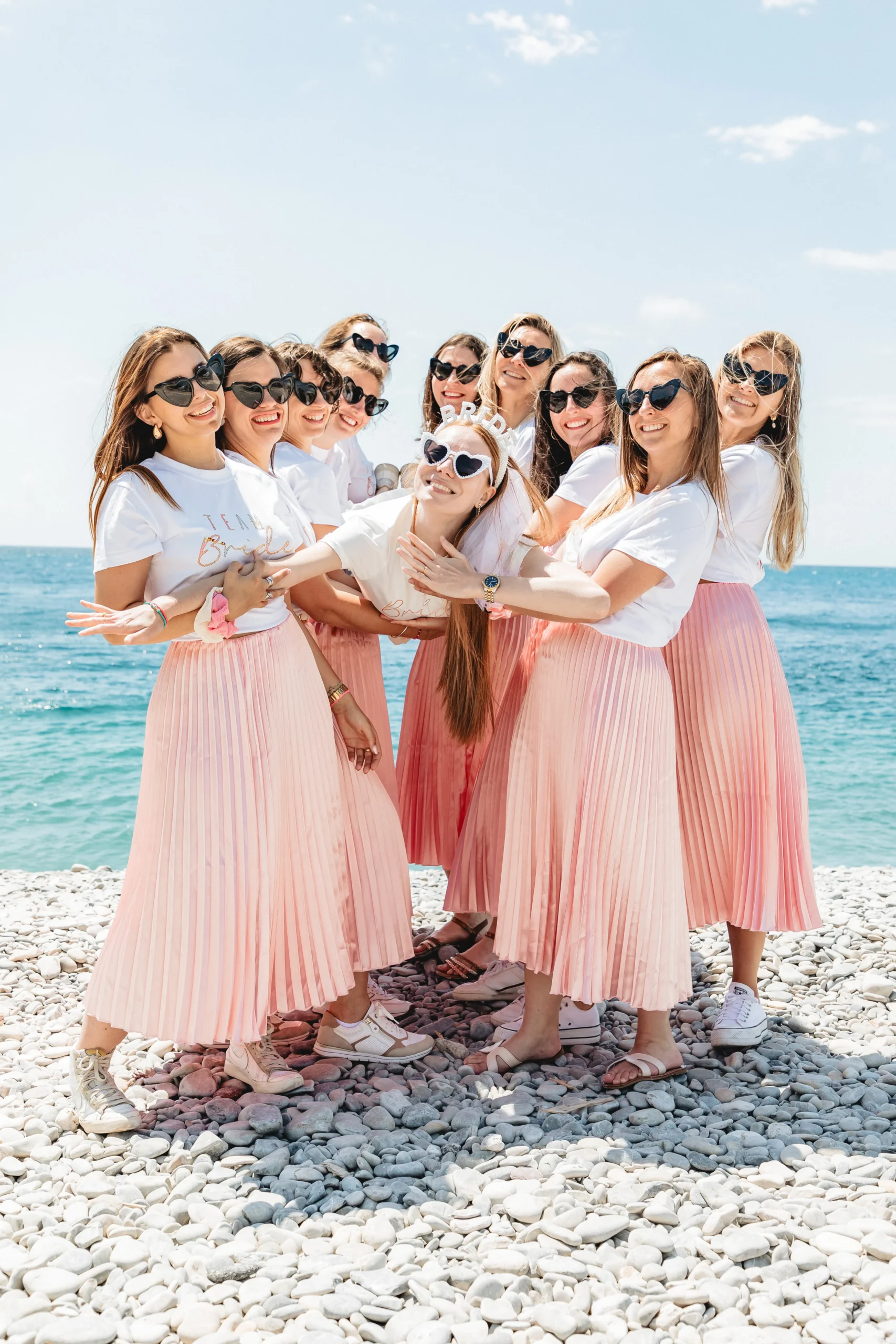 EVJF à Nice – Les amies soulèvent joyeusement la mariée au-dessus des galets avec la mer en toile de fond