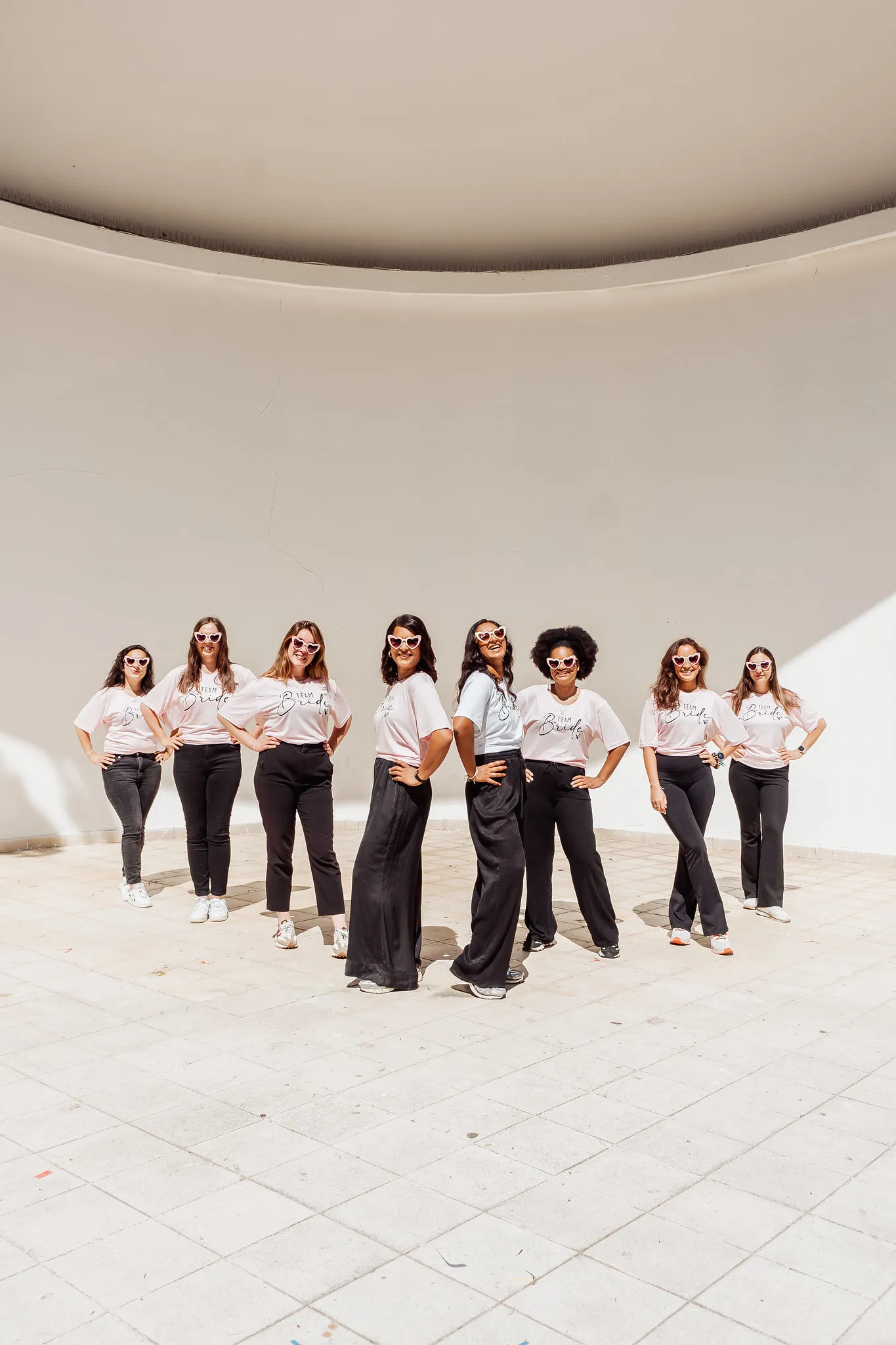 EVJF à Porto – Photo de groupe stylée et coordonnée en plein soleil Un groupe de femmes posant en formation, vêtues de tenues assorties “Team Bride”, lunettes en cœur, dans un espace architectural lumineux à Porto.