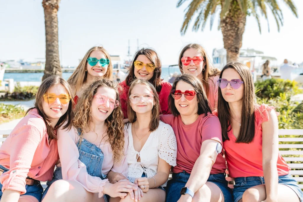 Photographe EVJF Split - Shooting EVJF Split Groupe EVJF à Split en lunettes colorées posé sur un banc sous les palmiers, ambiance bord de mer détendue.