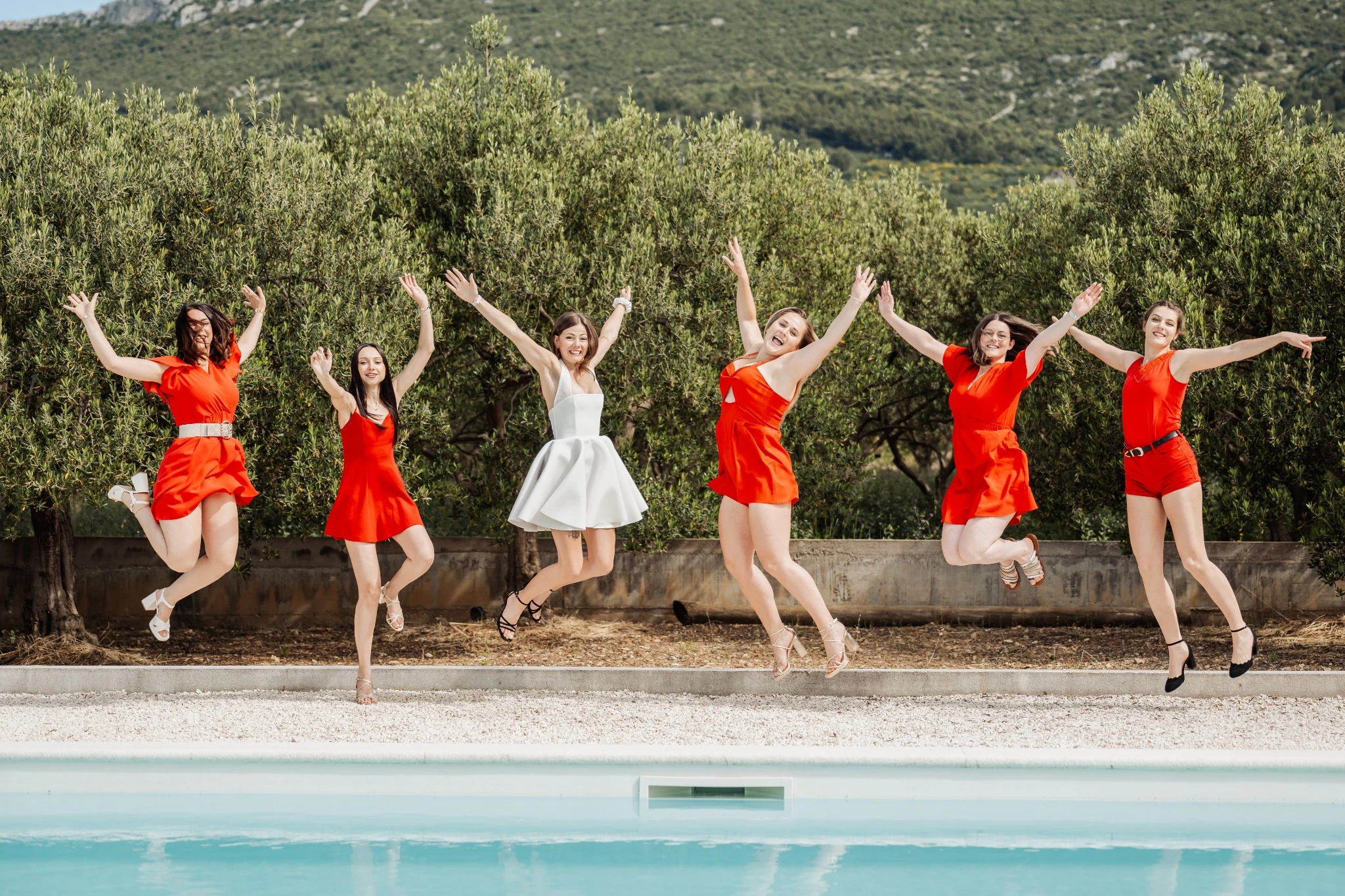 Groupe d’amies sautant en l’air lors d’un EVJF à Split, avec la mariée en robe blanche et les filles en rouge devant des oliviers.