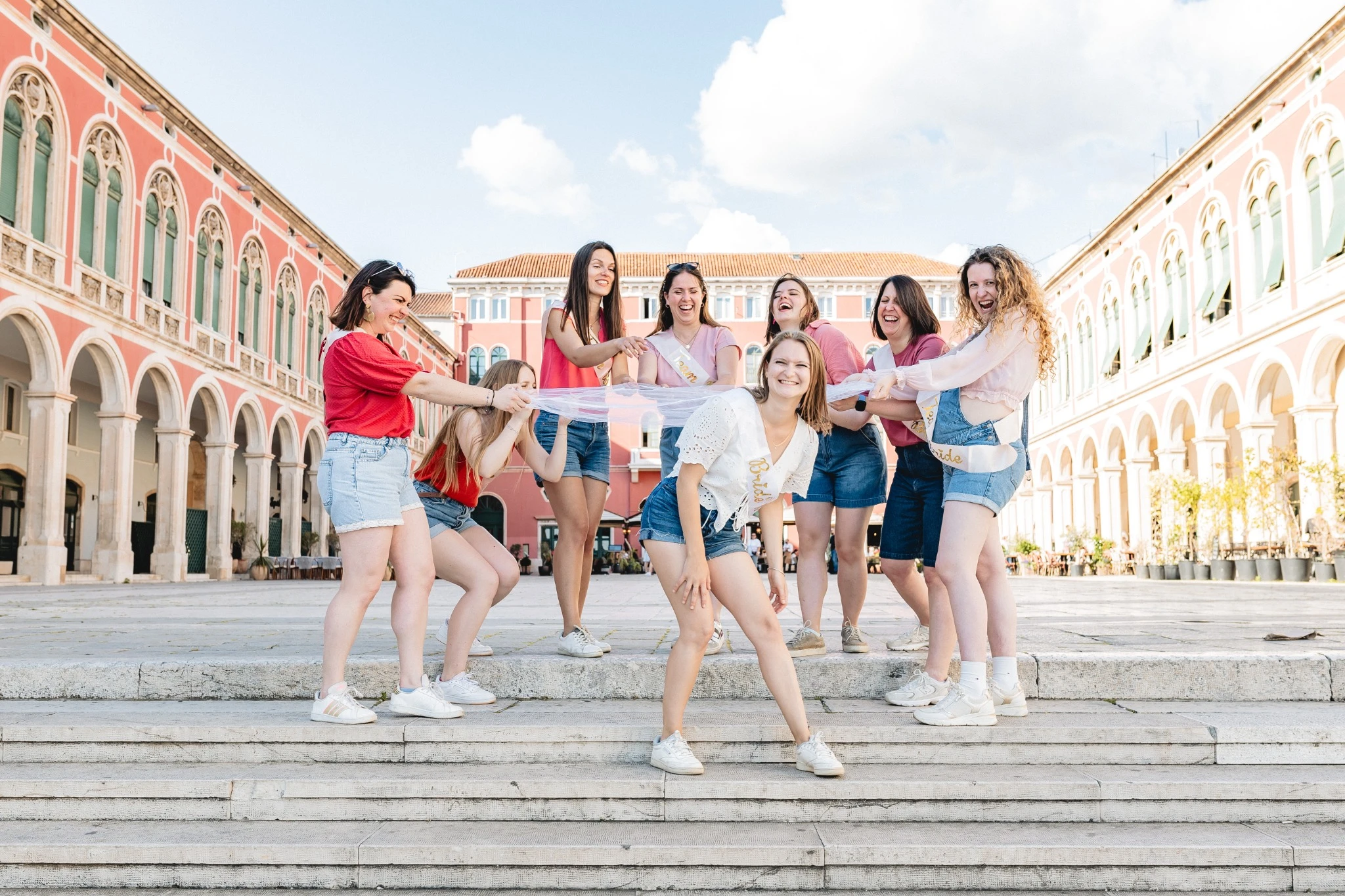 Groupe de filles en tenue estivale tirant le voile de la future mariée sur la place Prokurative à Split lors d’un EVJF plein de bonne humeur.