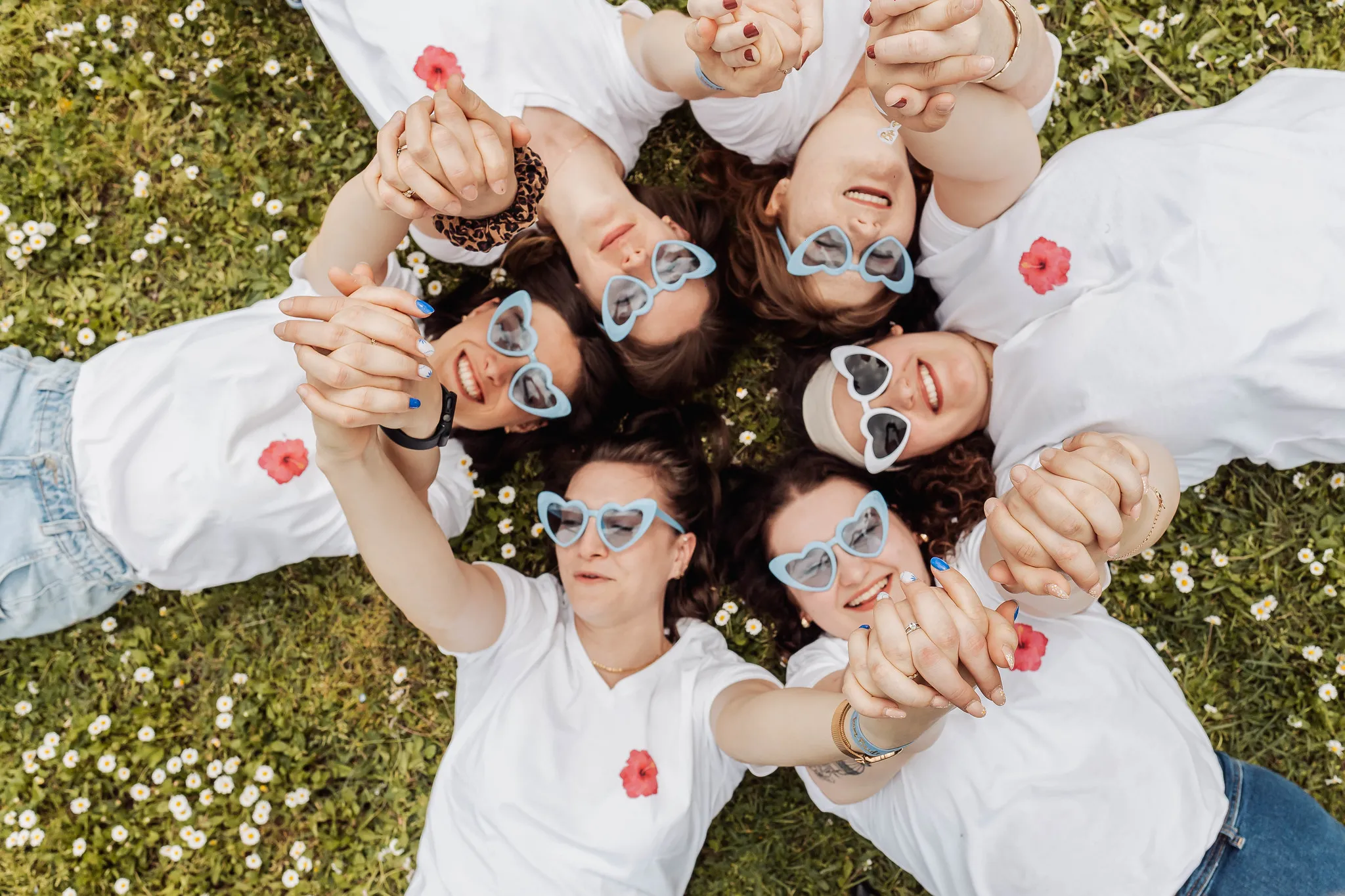 EVJF à Toulon – Amies allongées en cercle dans un champ fleuri Groupe de femmes allongées dans l’herbe, en cercle, portant des lunettes cœur bleues et se tenant les mains pendant un EVJF à Toulon.