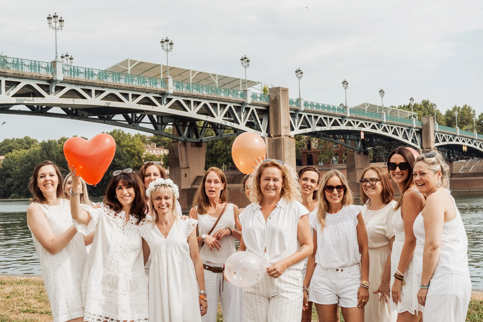 Groupe de femmes en blanc célébrant un EVJF au bord de la Garonne à Toulouse, tenant des ballons colorés devant le pont Saint-Pierre.