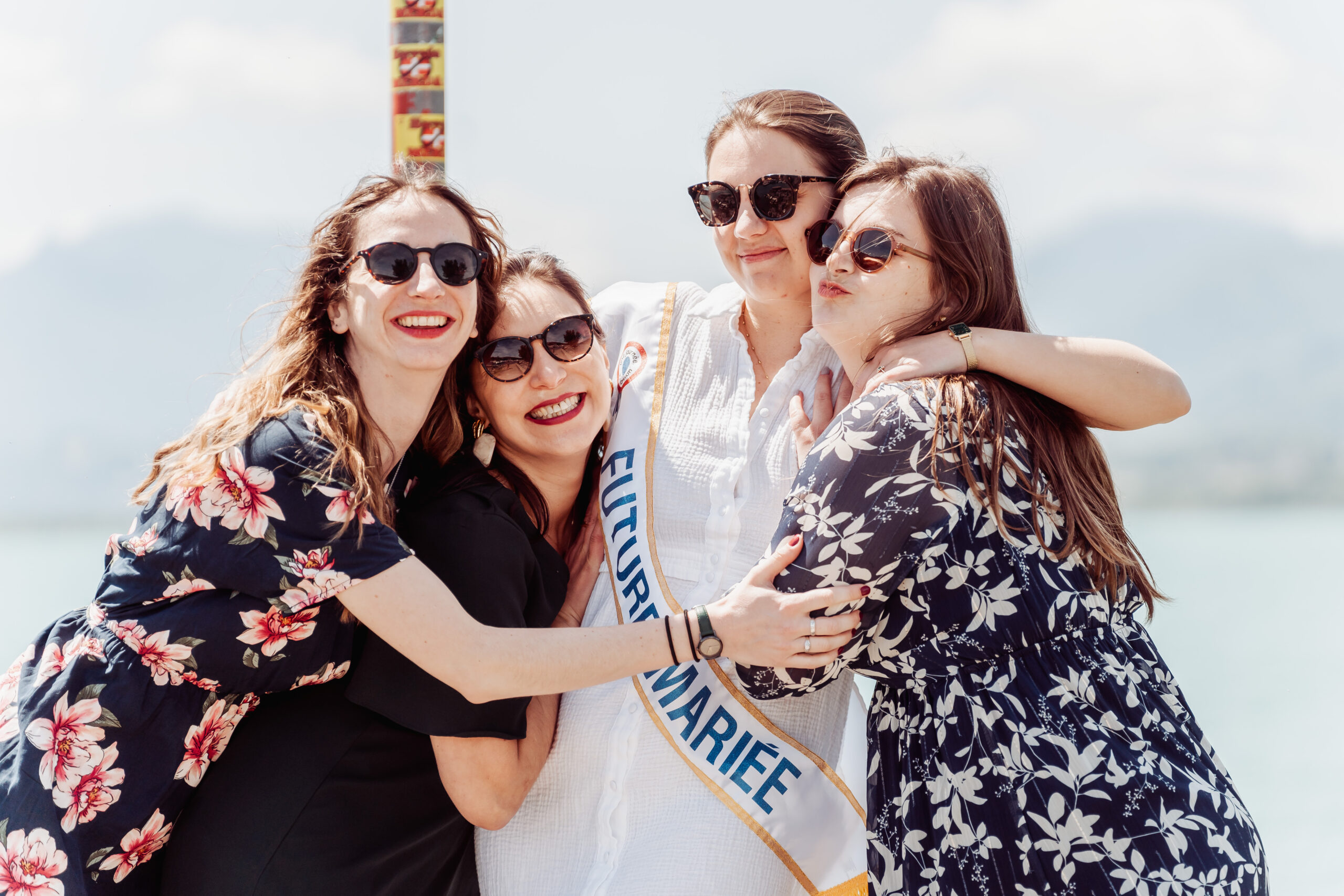 Photo de groupe lors d’un EVJF à Aix-les-Bains, quatre femmes enlacées au bord du lac avec la future mariée portant une écharpe “futur·e marié·e”, dans une ambiance joyeuse et complice face aux montagnes.