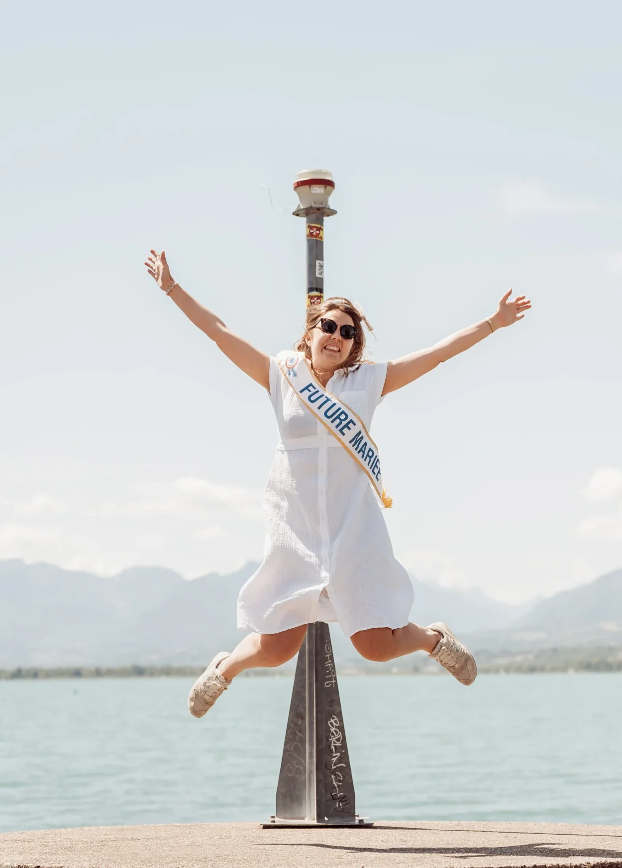 Saut de la future mariée lors d’un EVJF à Aix-les-Bains au bord du lac, avec vue sur les montagnes, en robe blanche et écharpe “Future Mariée”