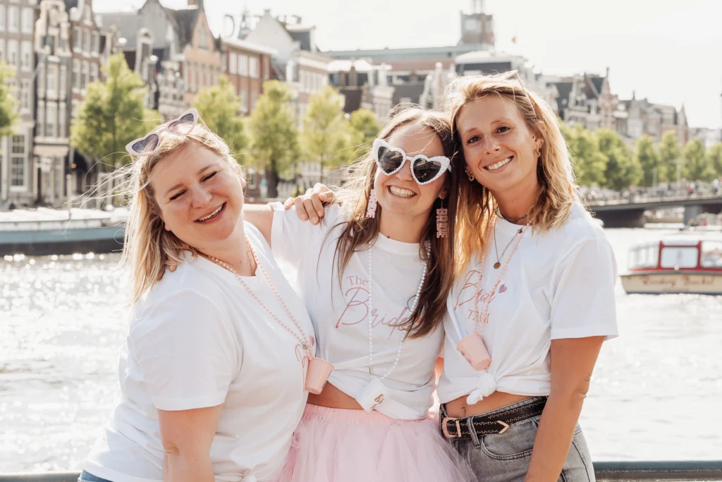 shooting photo evjf amsterdam Trois femmes posant souriantes lors d’un enterrement de vie de jeune fille à Amsterdam, habillées en blanc avec des lunettes fantaisie en forme de cœur, devant un canal typique et des maisons néerlandaises.