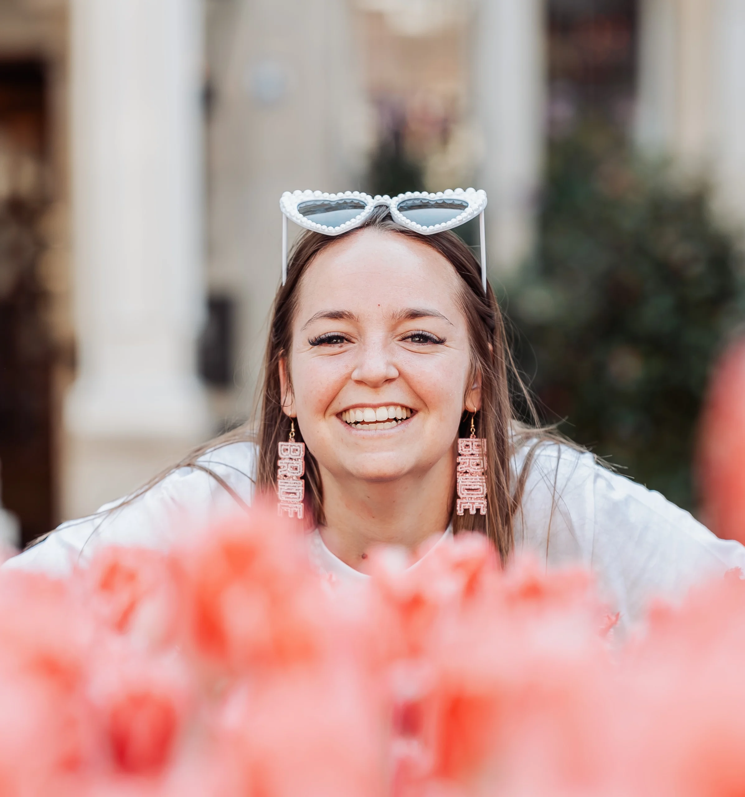 portrait-evjf-amsterdam-mariee-fleurs-roses Portrait en gros plan d’une future mariée souriante lors d’un EVJF à Amsterdam, portant des lunettes fantaisie et des boucles d’oreilles "bride", photographiée derrière un champ de fleurs roses.