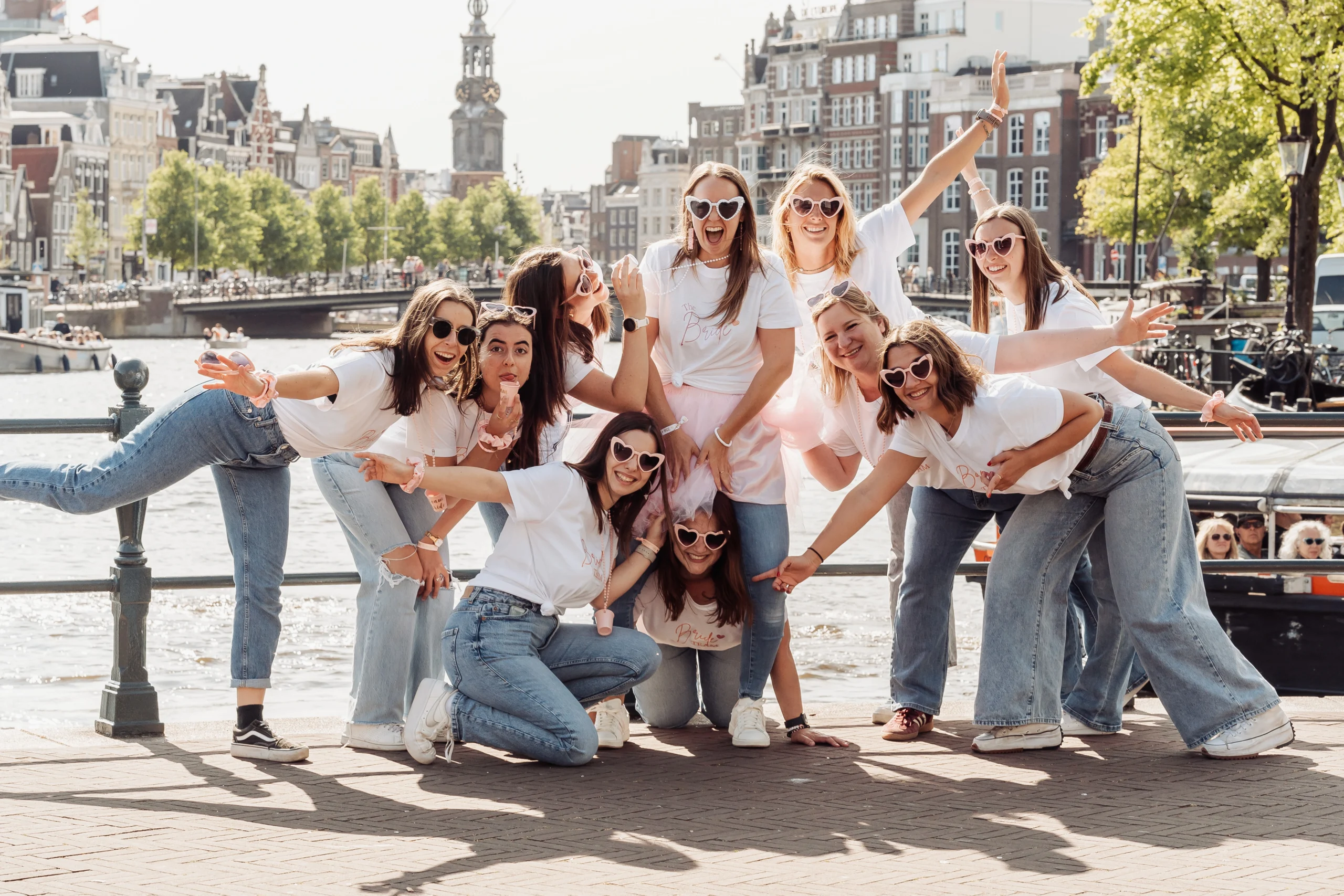 team-bride-amsterdam-style-denim-canal-pose Photo de groupe lors d’un EVJF à Amsterdam : une dizaine de femmes en jeans et tee-shirts blancs, portant des lunettes fantaisie, posent avec énergie au bord d’un canal typique de la ville, avec des bateaux et bâtiments hollandais en arrière-plan